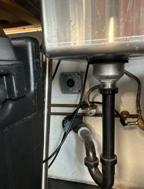 Under-sink view of a stainless steel sink with plumbing and a wall-mounted gray box with wiring.