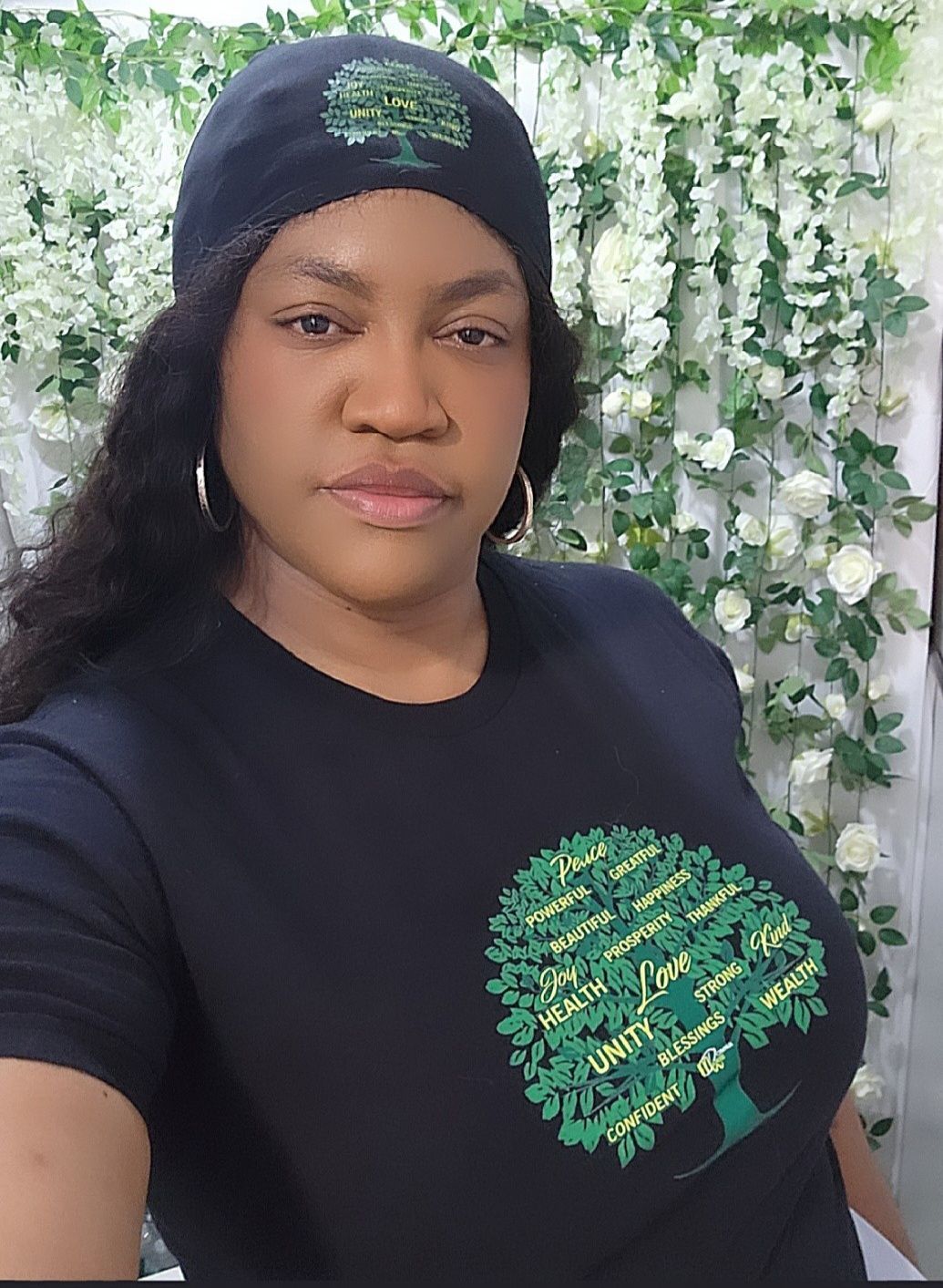 A woman wearing a black t-shirt with a tree on it is standing in front of a wall of flowers.