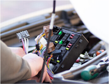 Hands working on a car's fuse box, with colorful wires and fuses visible. | All Valley Car Care