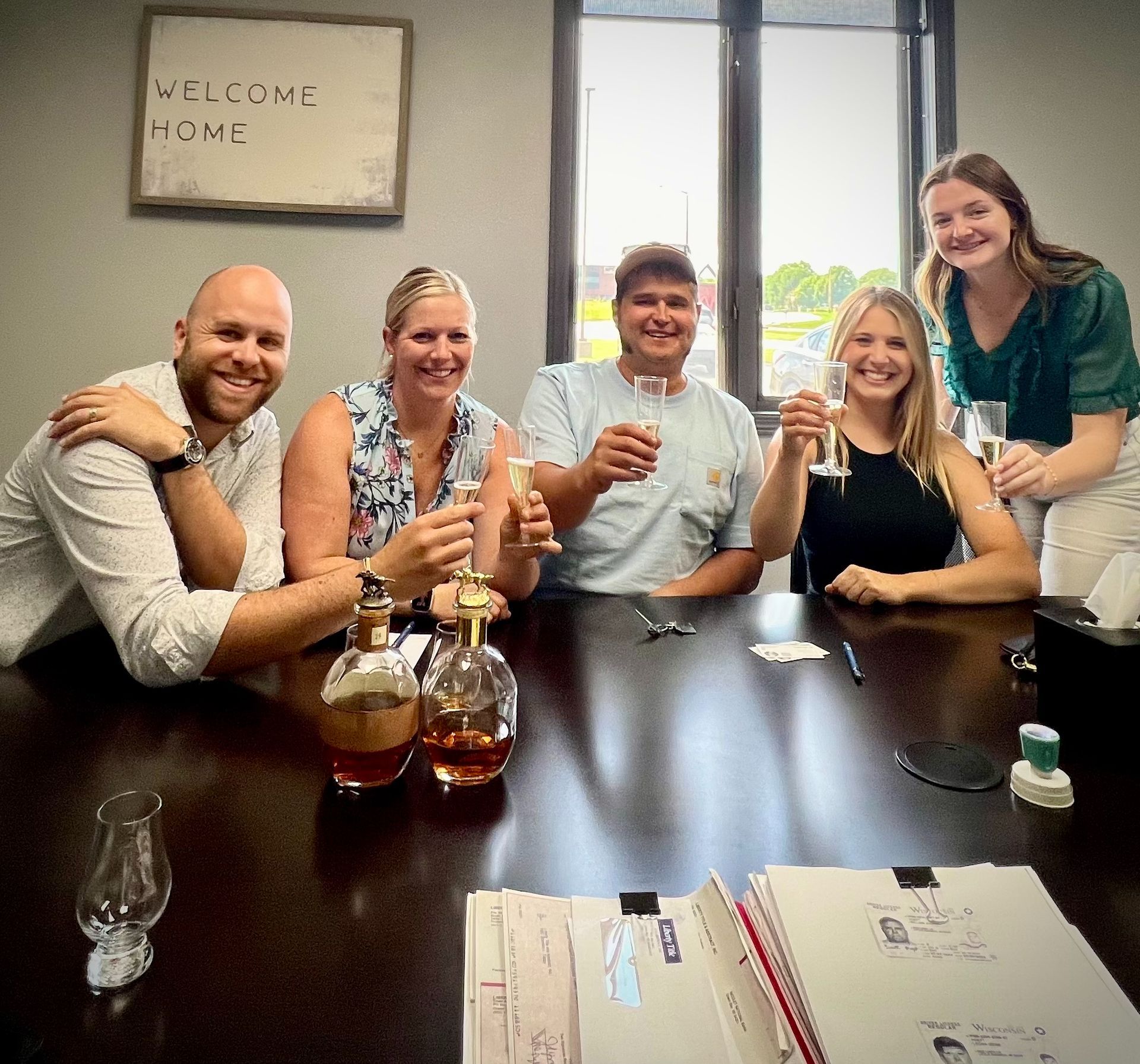 A group of people are sitting around a table with bottles of alcohol and a sign that says welcome home