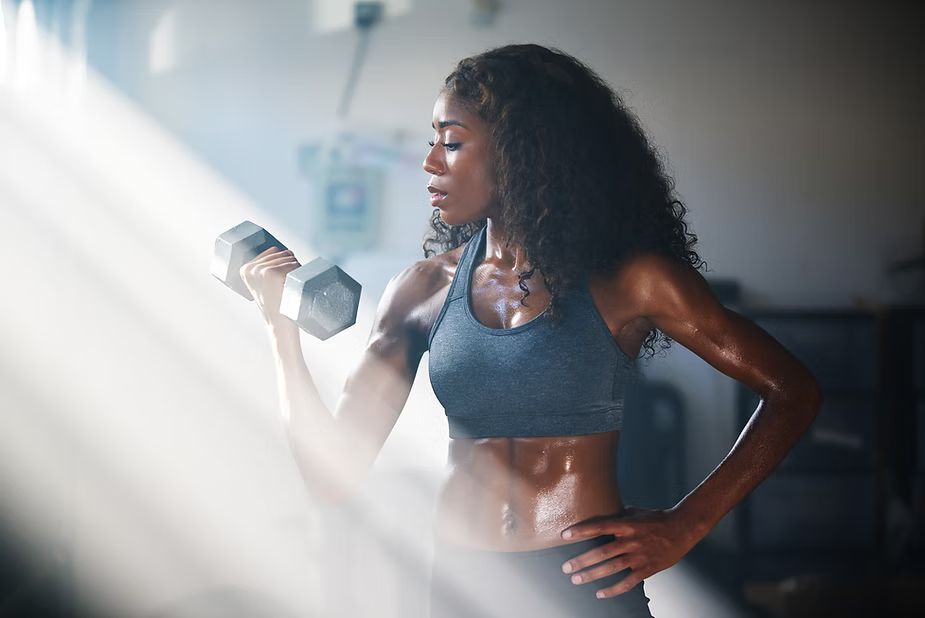 Woman lifting a dumbbell in a home gym, sweating, wearing a sports bra and leggings.