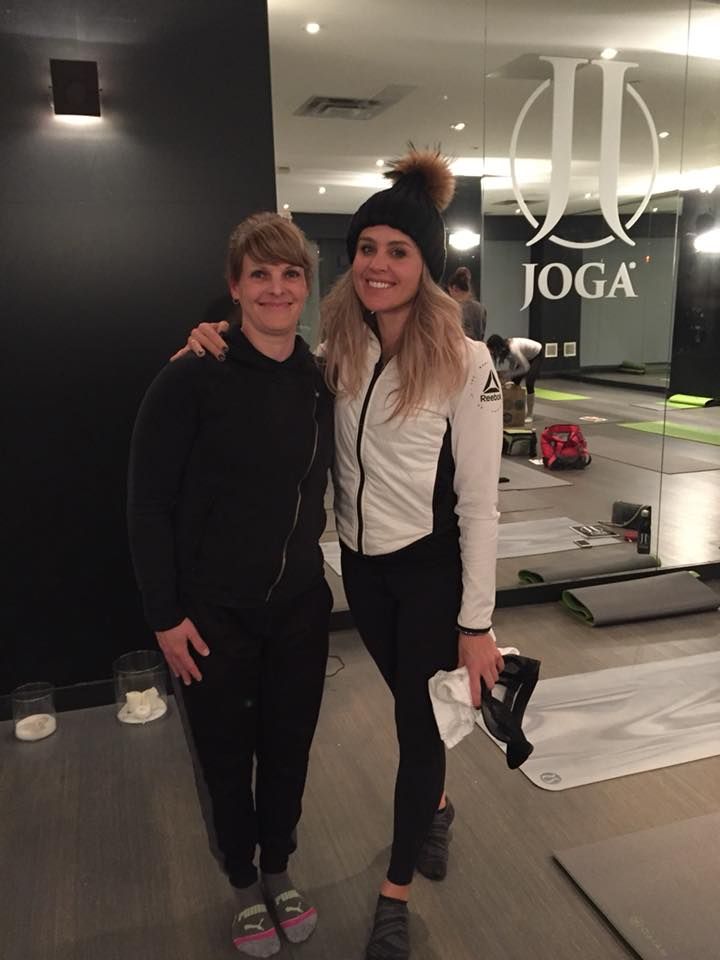 Two women pose in a yoga studio. One wears a black outfit, the other a white jacket and black beanie.