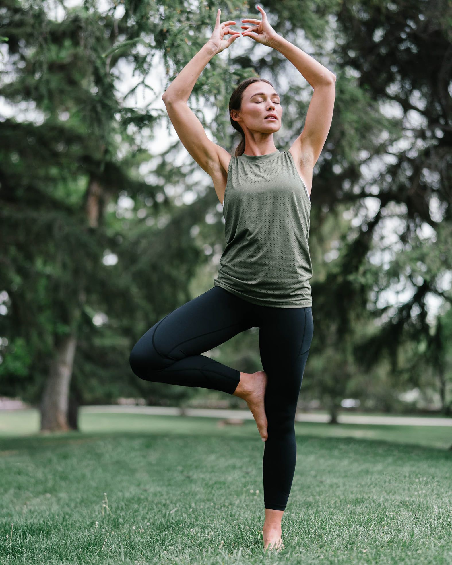 Woman in tree pose outdoors with arms raised, eyes closed, wearing green tank top and black leggings.