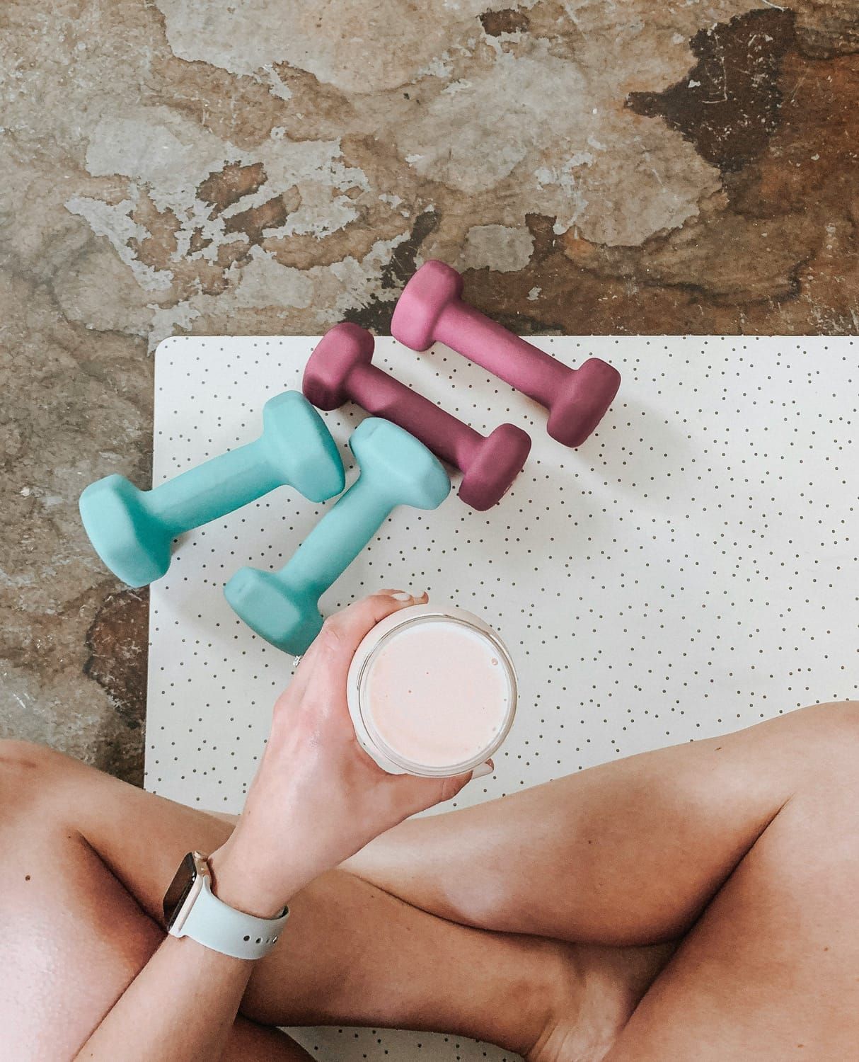 Woman sitting with smoothie, weights, and watch on a yoga mat.