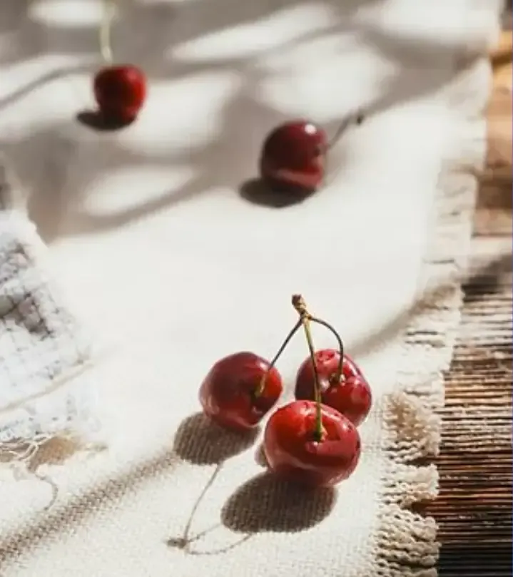 Cherries on a white, textured cloth, with shadows. Sunlight and a rustic wooden background.