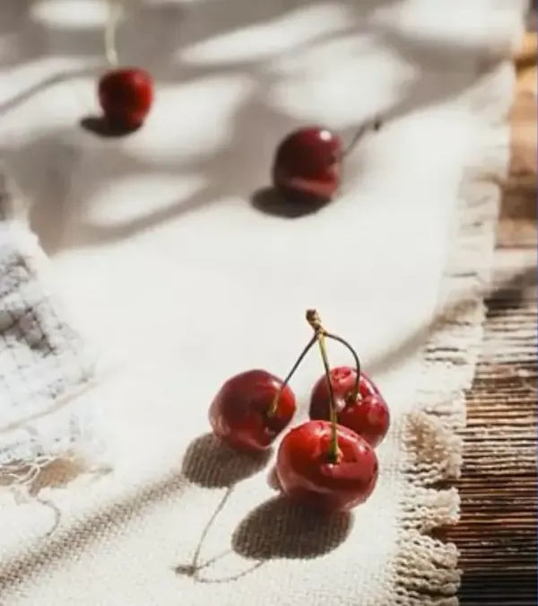 Cherries on a white, textured cloth, with shadows. Sunlight and a rustic wooden background.