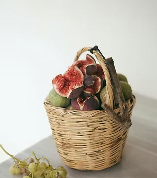 Wicker basket overflowing with figs, some halved, next to grapes; on a surface against a white background.