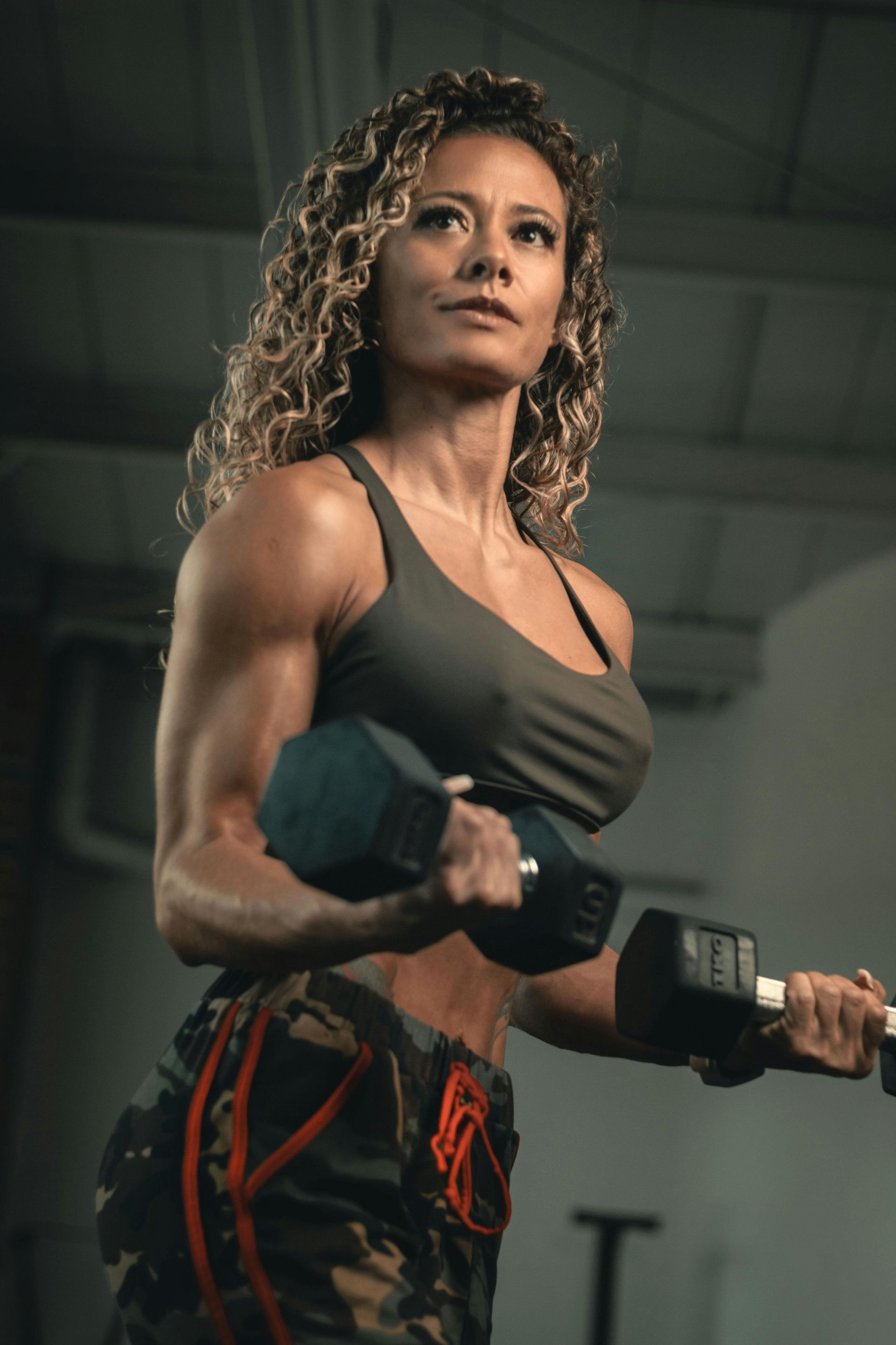 Woman with curly hair, lifting dumbbells, flexing arm muscles in a gym setting.