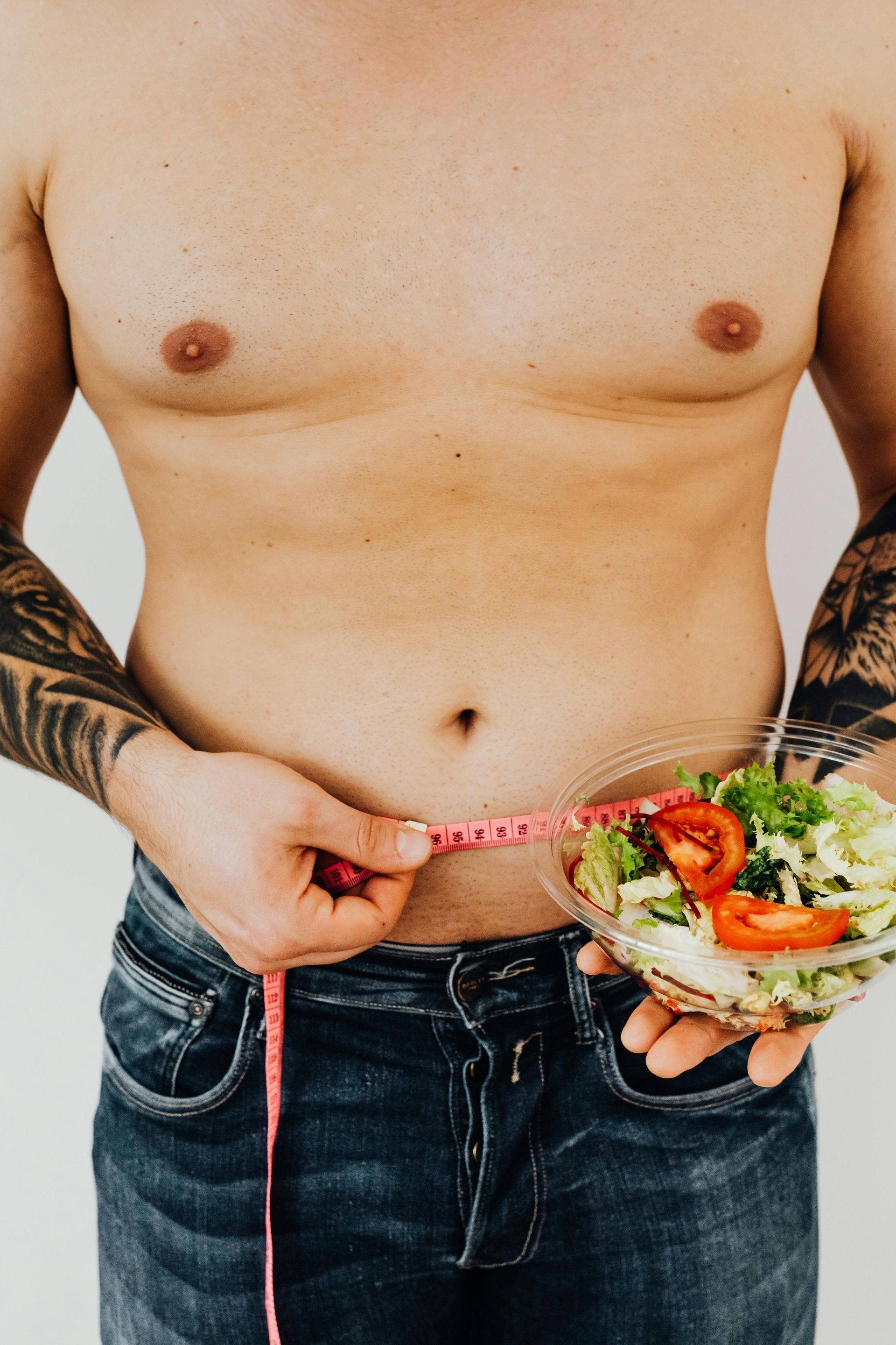 Shirtless man in jeans measures waist with a pink tape while holding a salad bowl.