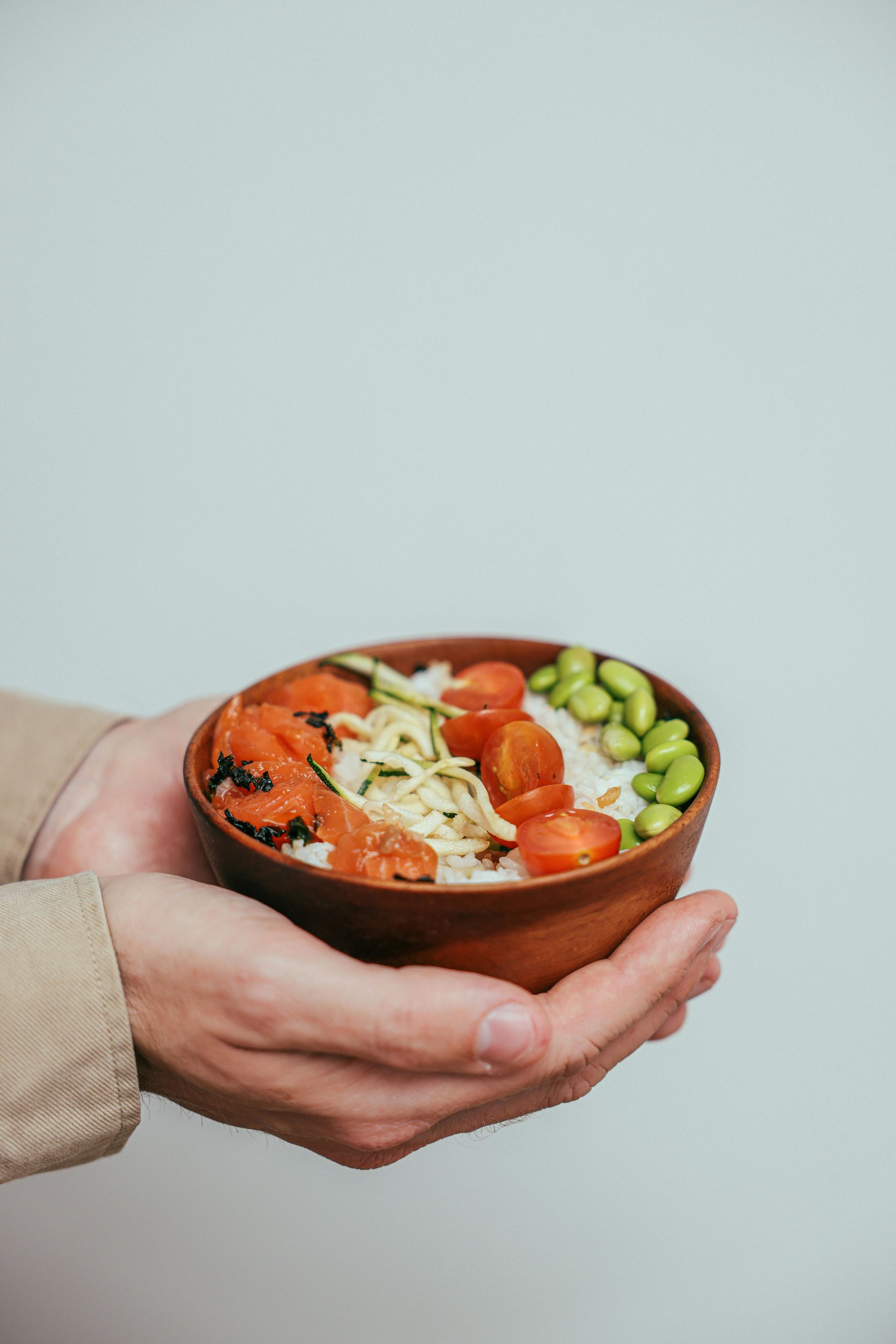 Person holding a wooden bowl filled with salmon, rice, tomatoes, edamame, and zucchini.