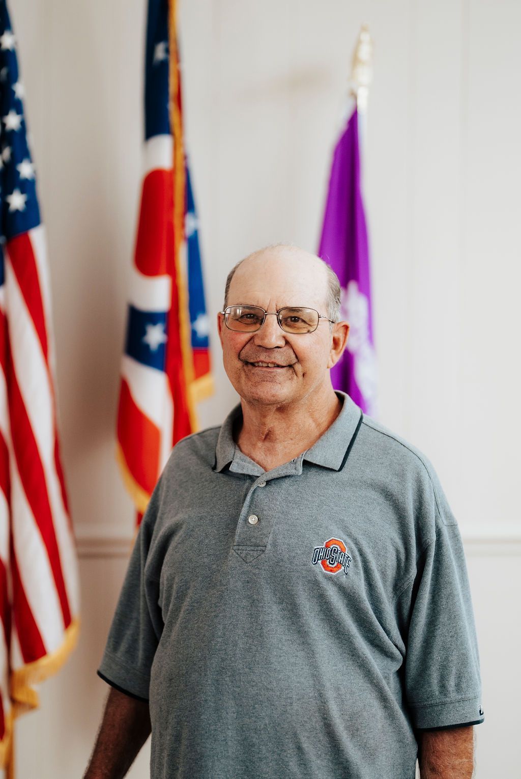 A man in a grey shirt is standing in front of two american flags.