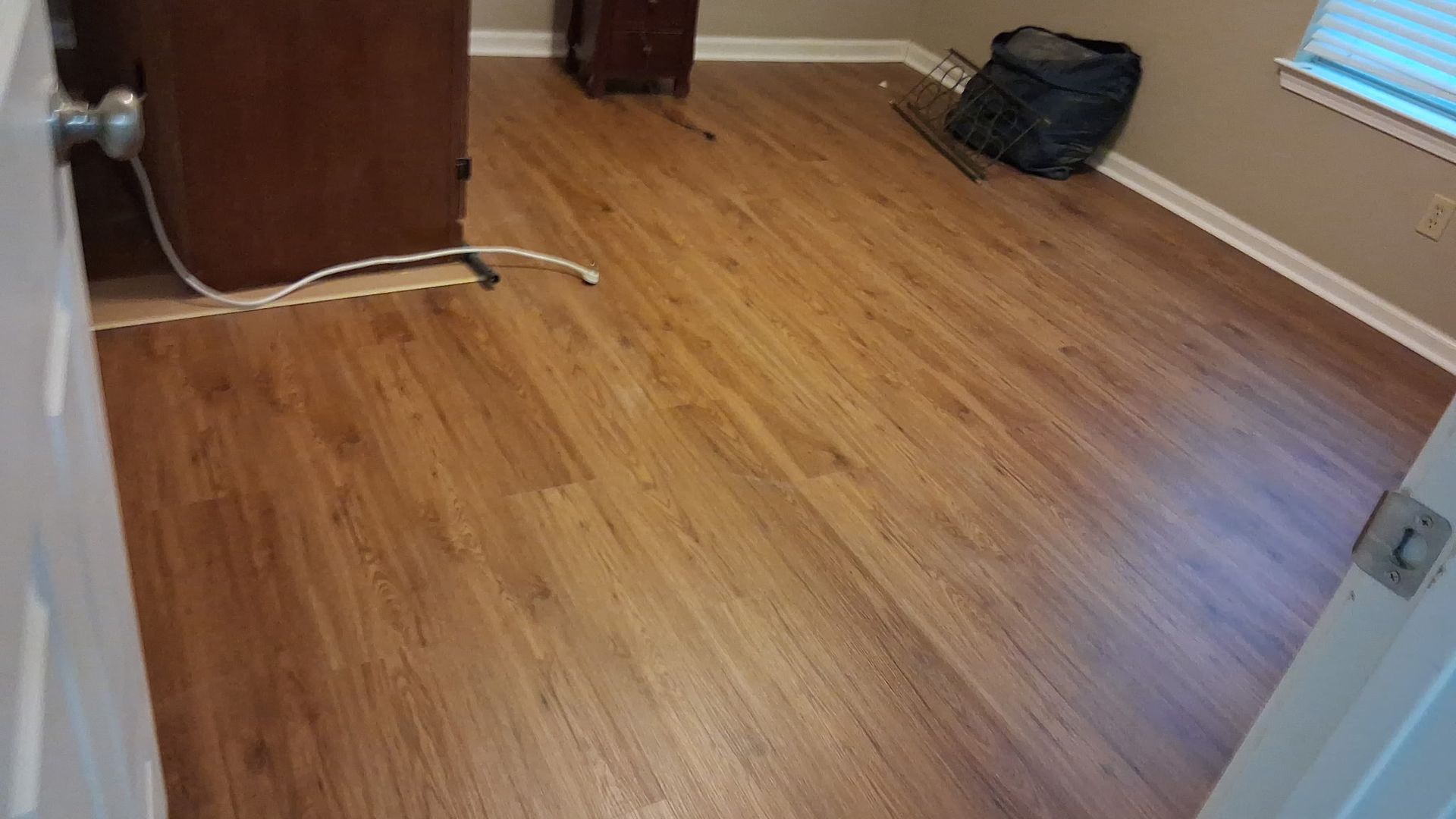Room with brown wood-look flooring, door frame in foreground, and furniture visible in background.