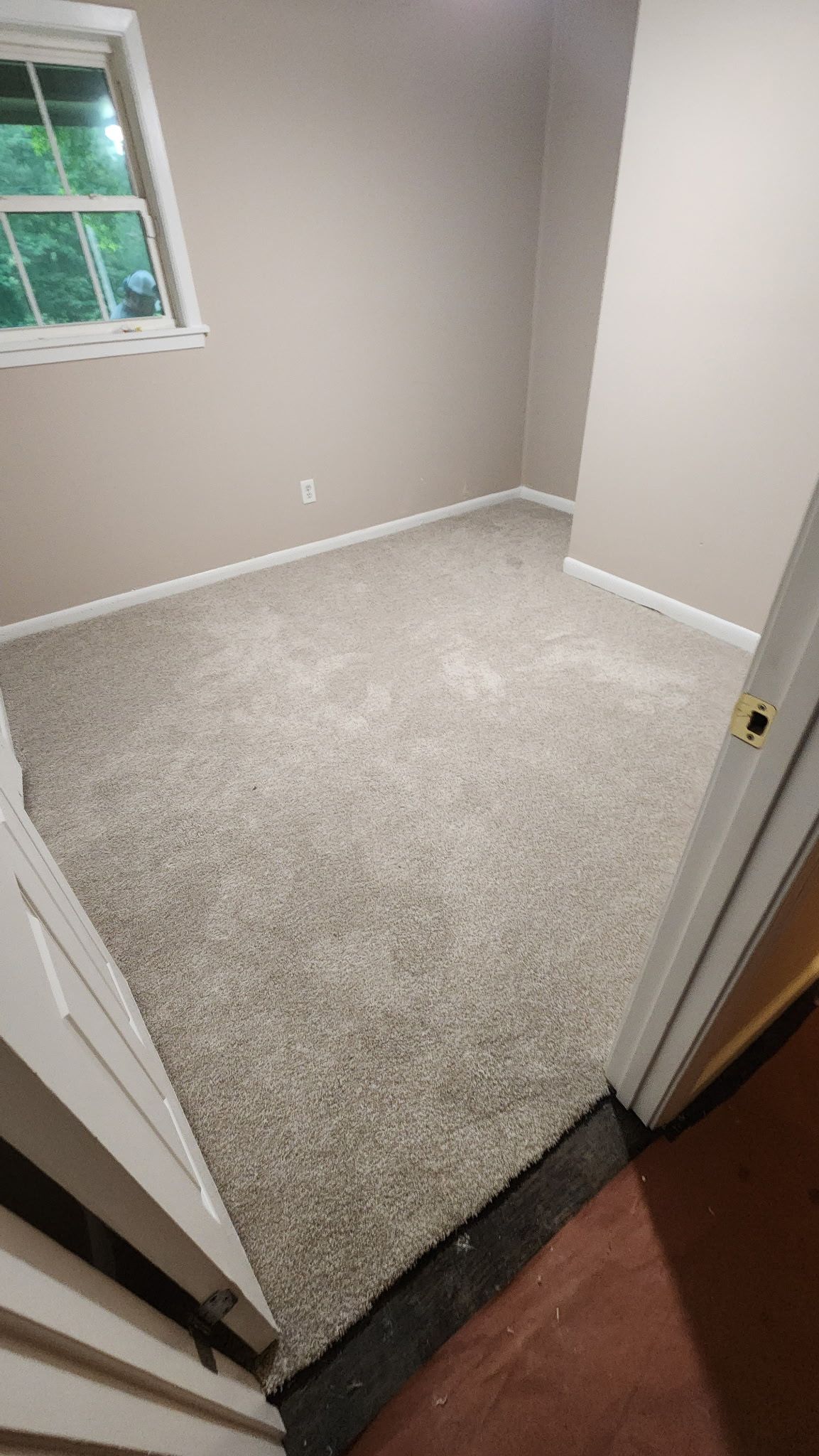 Small room with tan walls and light gray carpet, open doorway, window on the left.