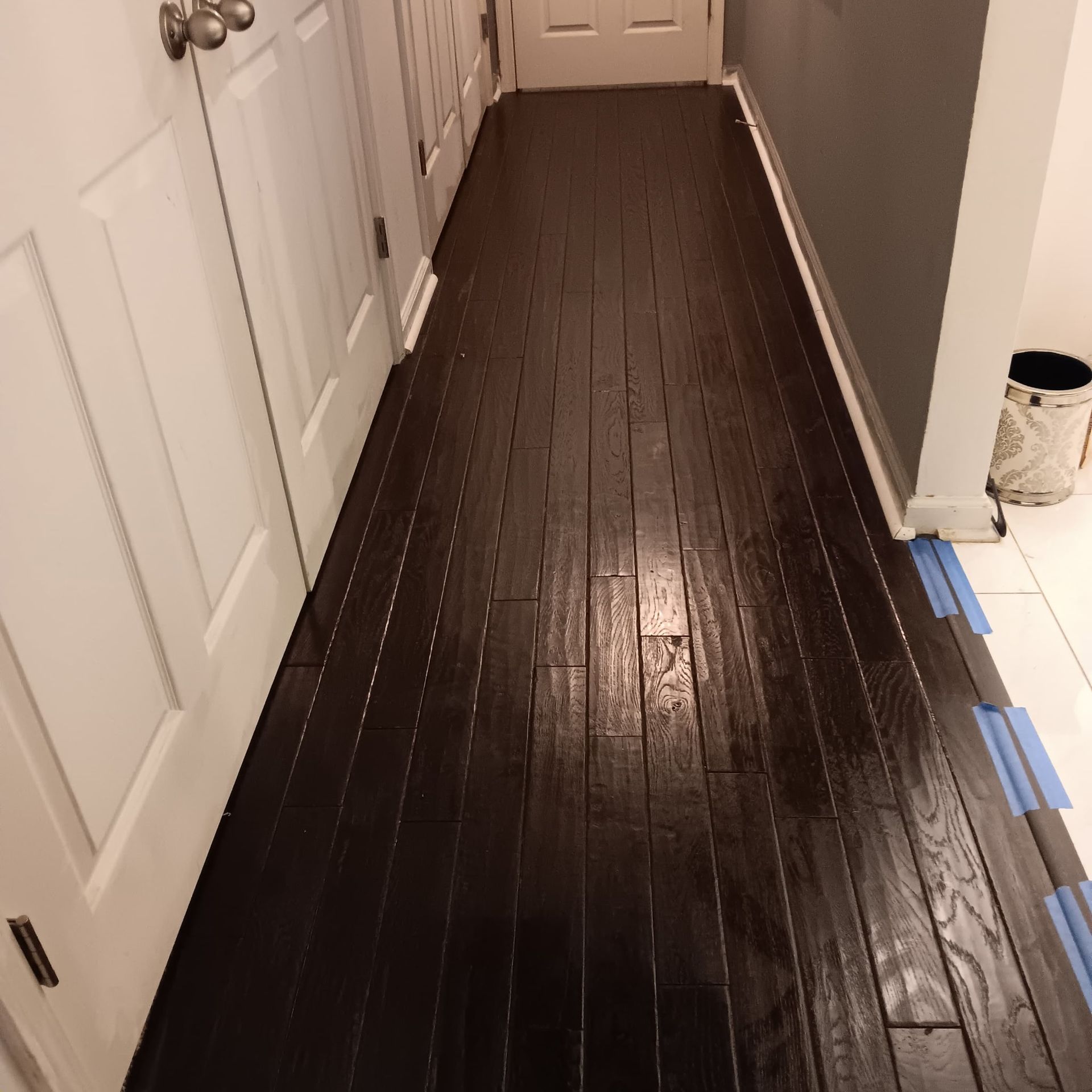 Dark stained wood floor in a hallway with white doors and a gray wall; a can and toilet are visible.