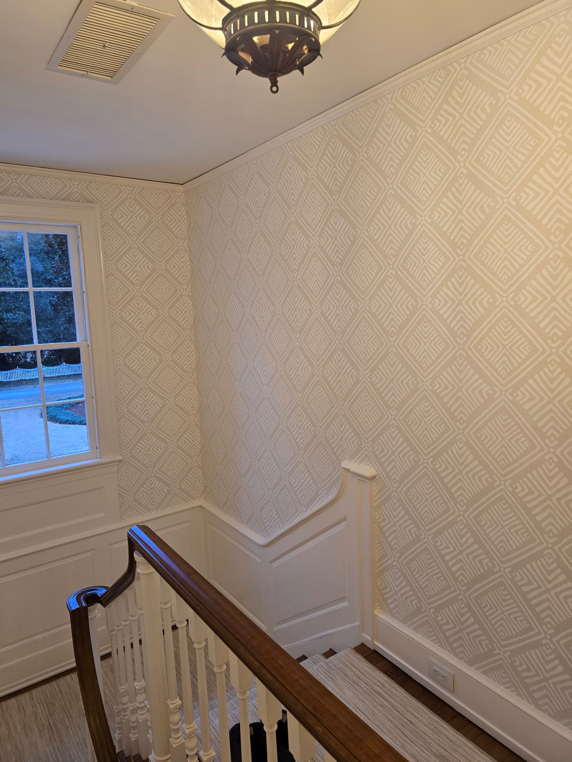 Staircase with light-colored patterned wallpaper, wooden banister, and a window overlooking a yard.