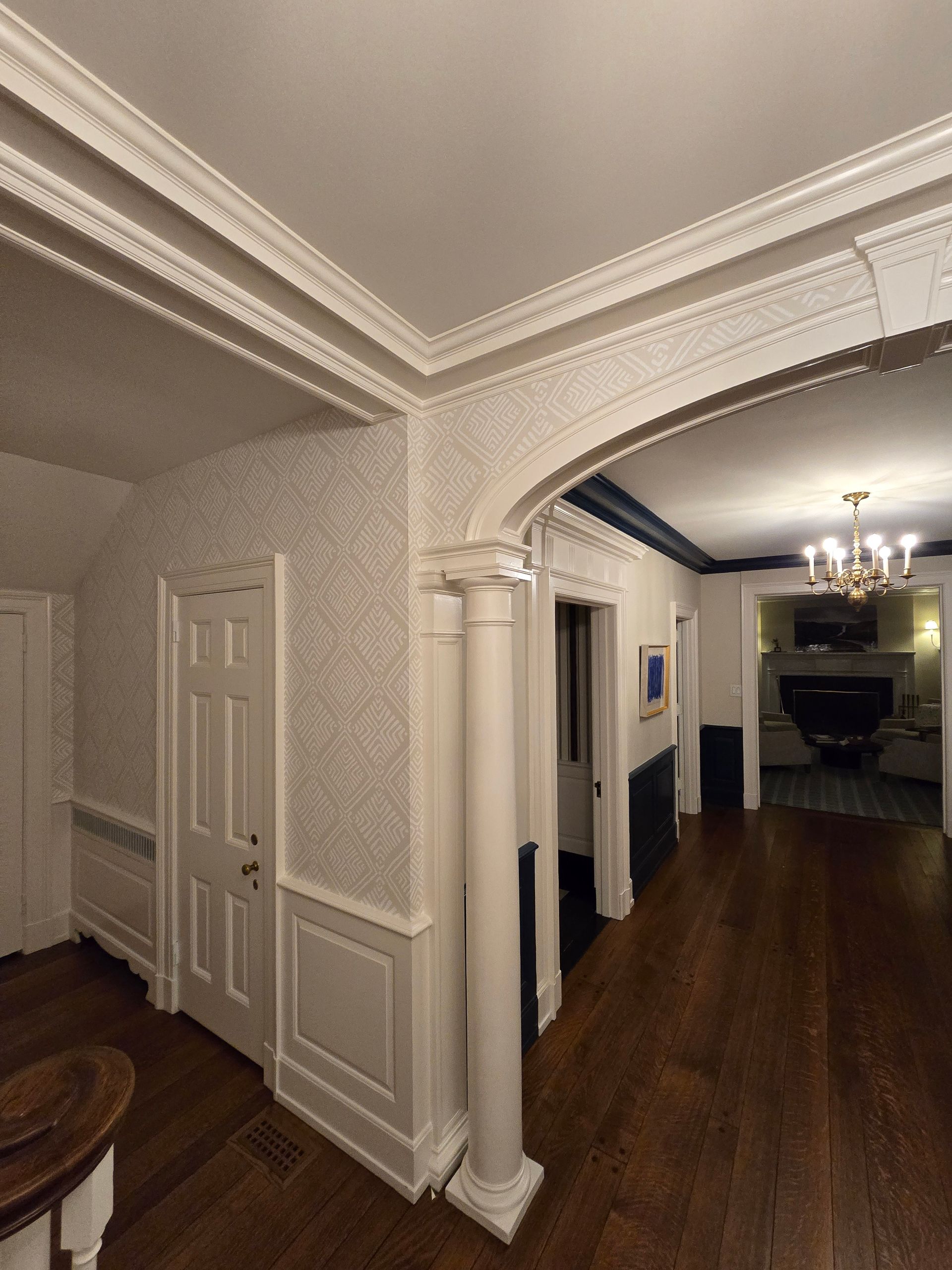 Hallway with arched doorway, white trim, and dark wood floor. A chandelier hangs in the distance.
