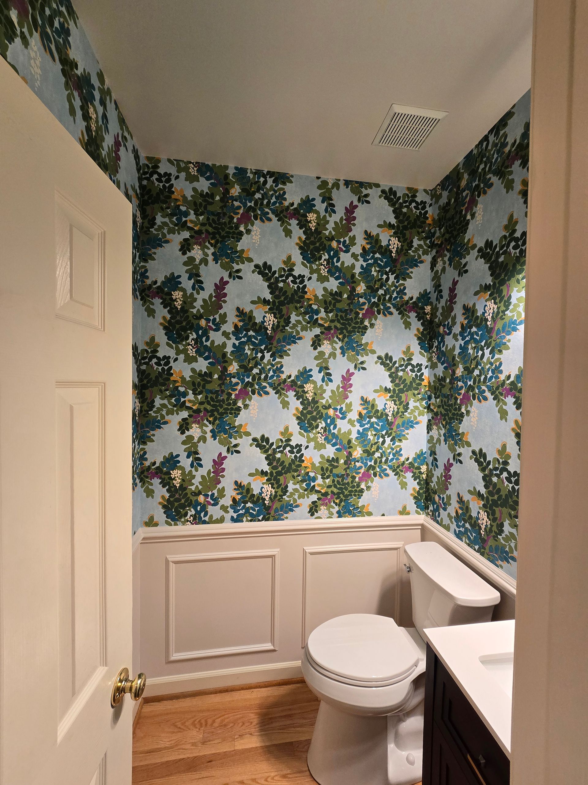 Small bathroom with floral wallpaper, white wainscoting, a toilet, and a partially visible sink.