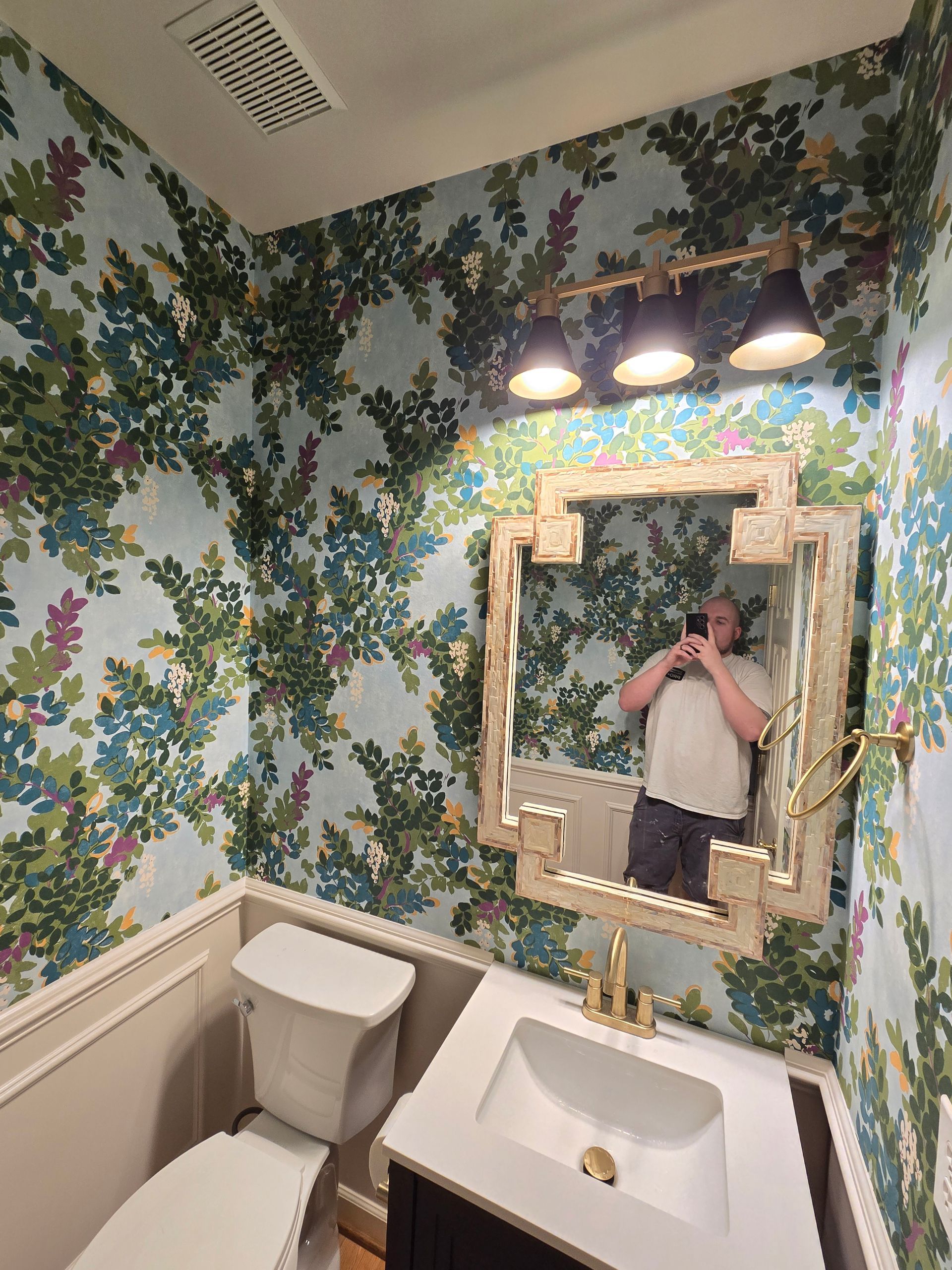 Bathroom with floral wallpaper, gold mirror and fixtures, and a person taking a photo in the mirror.