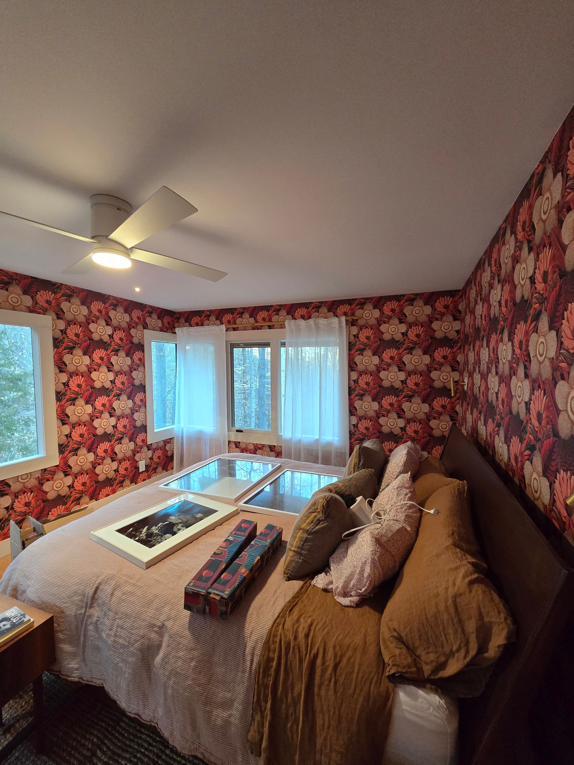 Bedroom with floral wallpaper, bed, windows with white curtains, and a ceiling fan.