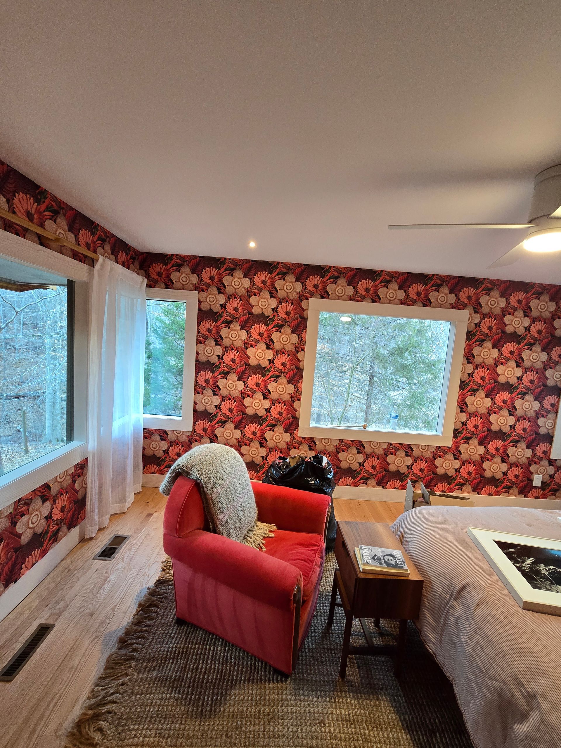 Cozy bedroom with red floral wallpaper, a red armchair, and large windows overlooking a green outdoor view.
