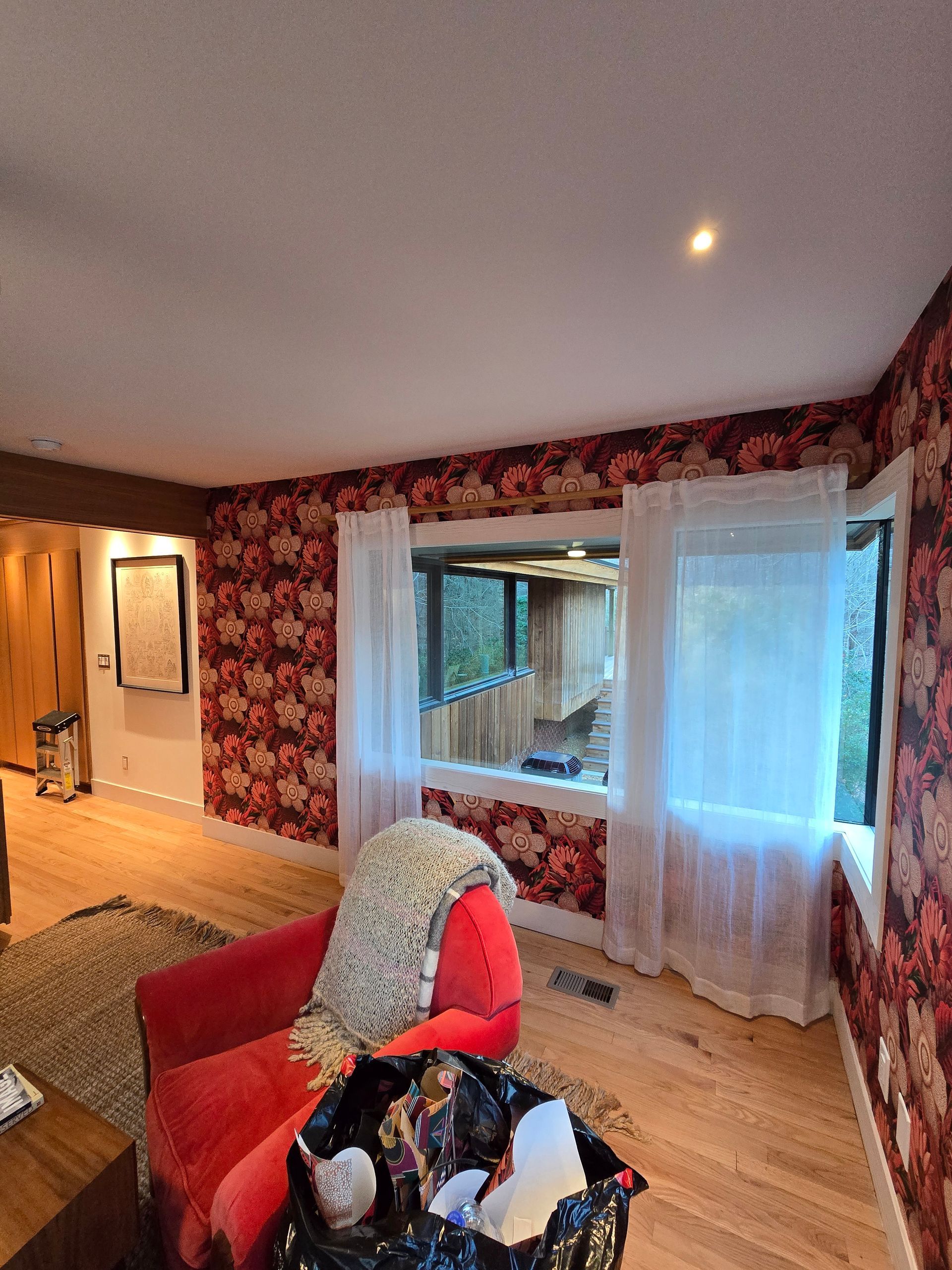 Cozy living room with floral wallpaper, red chair, sheer curtains, and a view of trees.