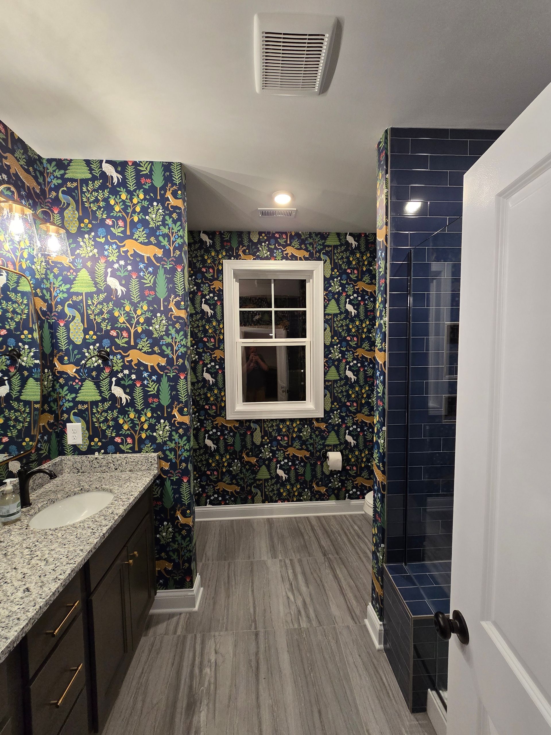 Bathroom with patterned wallpaper, dark cabinets, grey flooring, and blue tile shower.