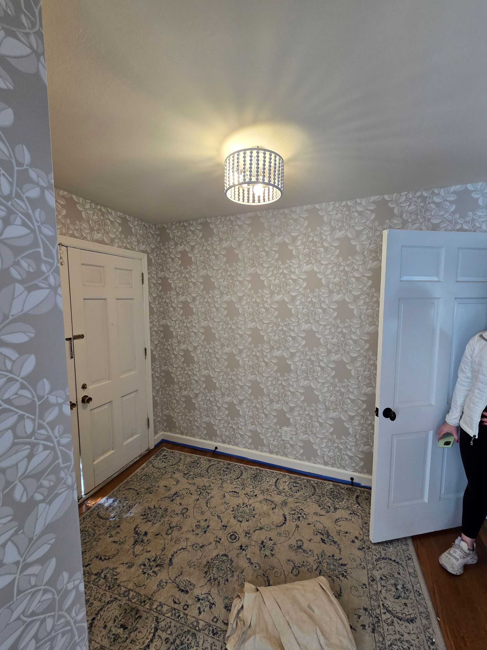Small entryway with floral wallpaper, white doors, and a chandelier. A person stands near a door.