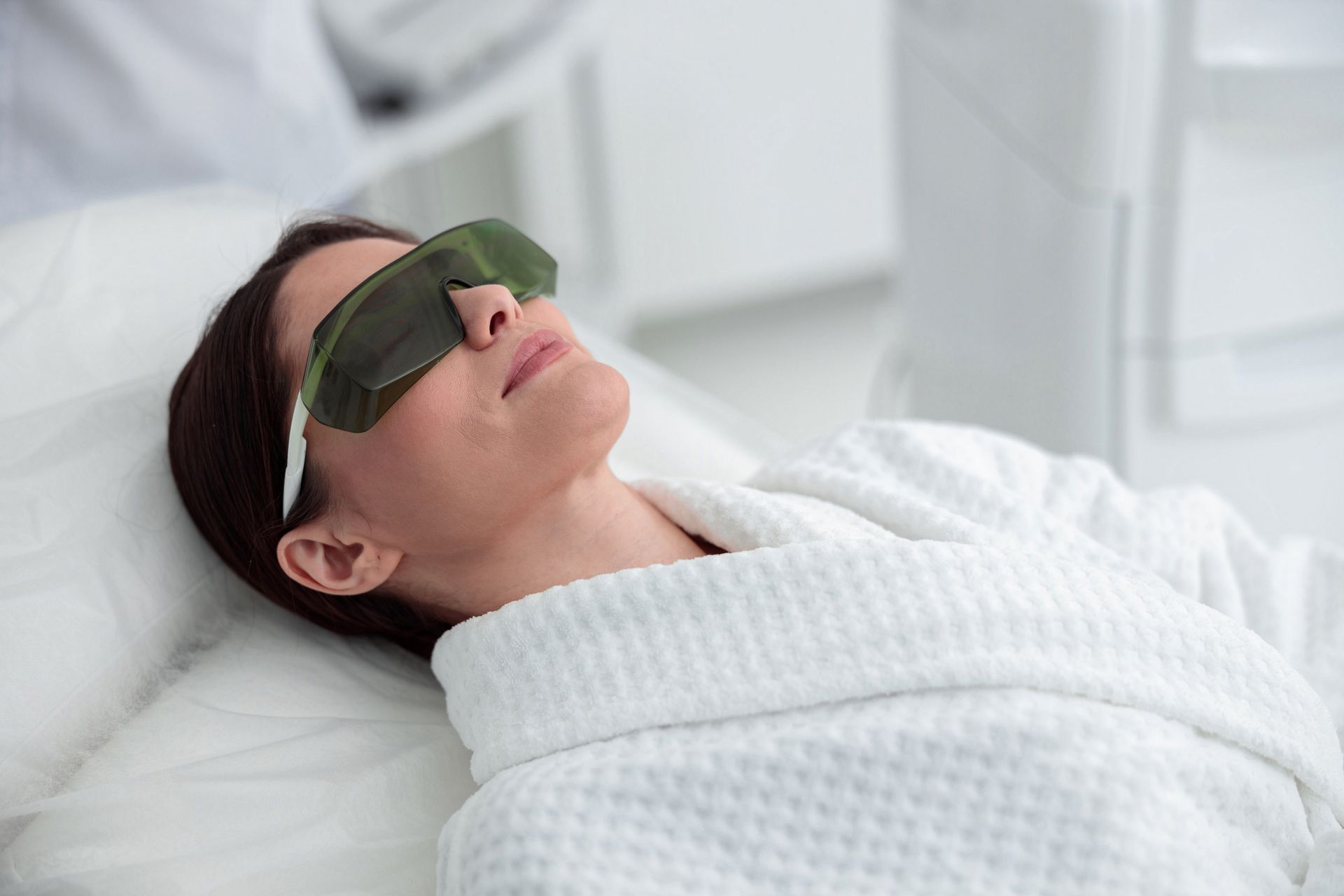 Woman wearing eye protection, lying on a treatment bed in a white robe, in a spa-like setting.