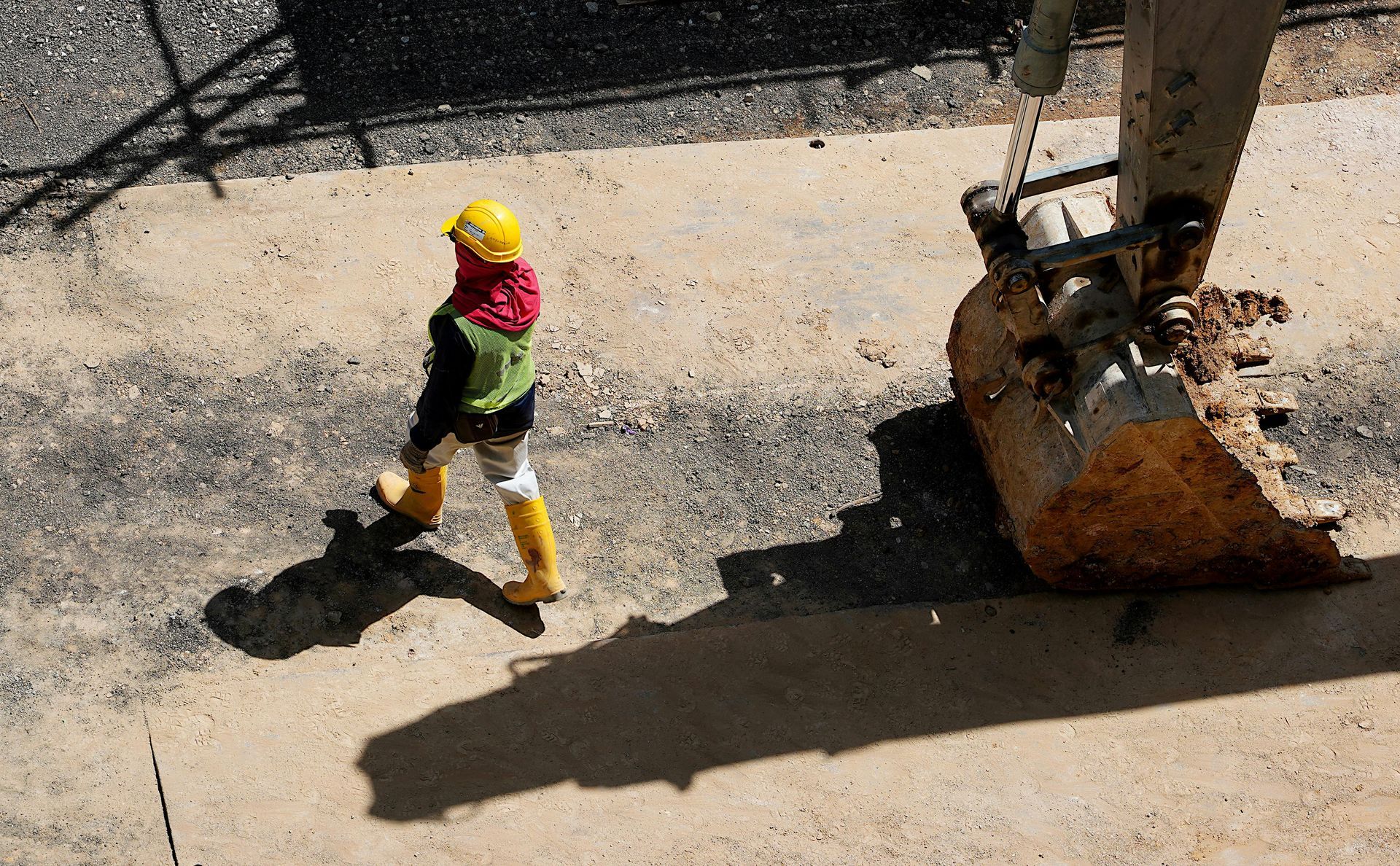 A man wearing a hard hat and yellow boots is walking in front of a large excavator bucket.