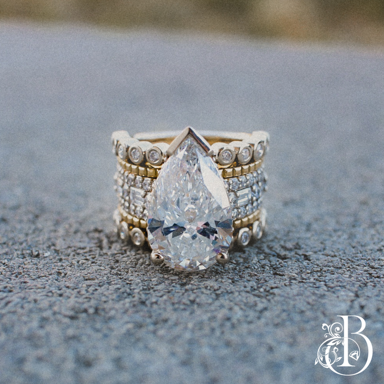 A pear-shaped diamond engagement ring with a yellow gold and diamond band, resting on a textured gray surface.