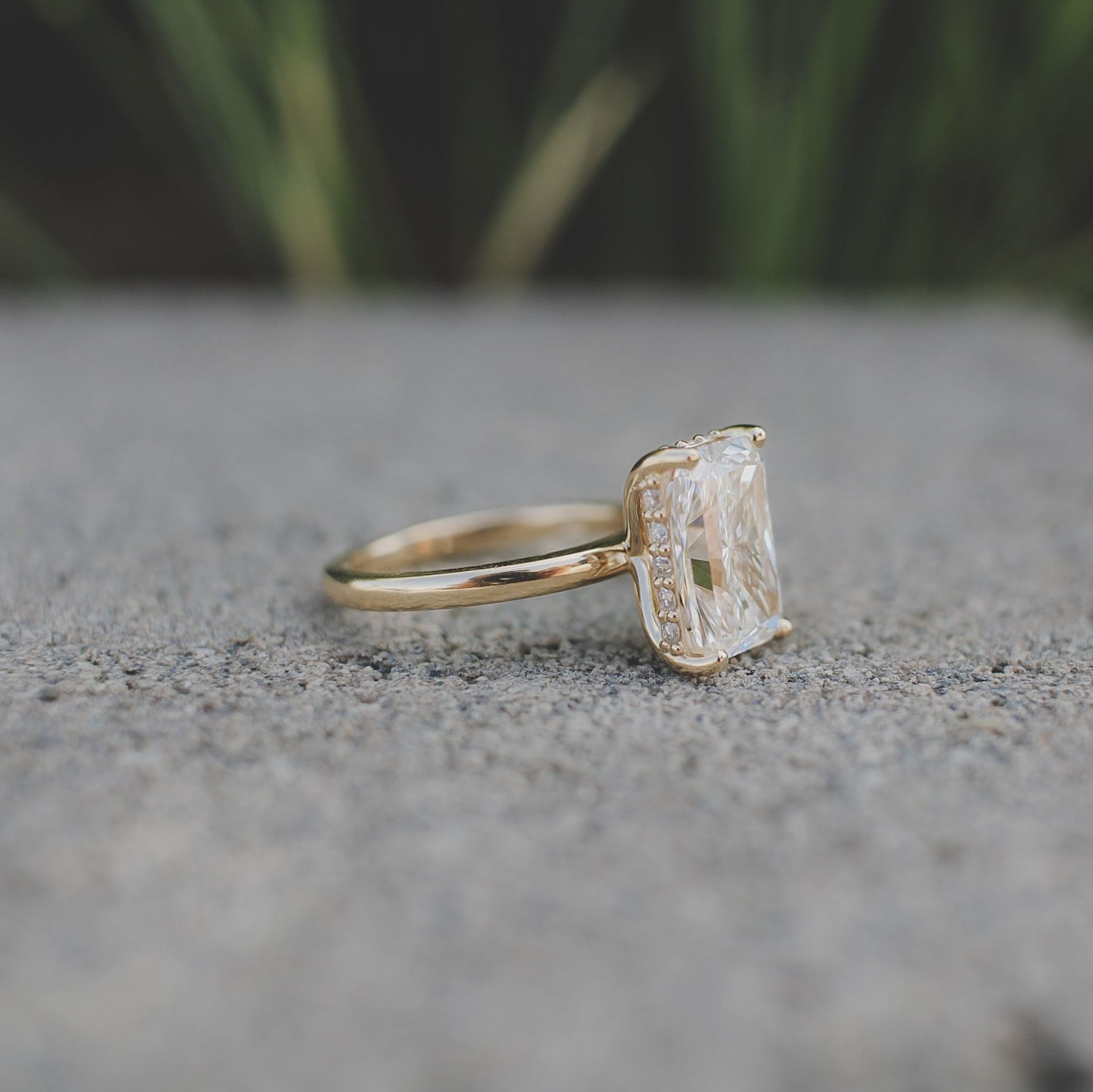 A gold engagement ring with a large rectangular-cut diamond, set against a blurred gray stone background.