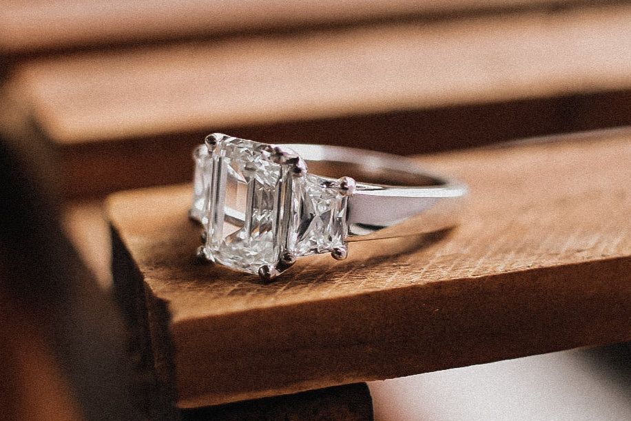 A three-stone engagement ring with a large emerald-cut center diamond resting on wooden slats.