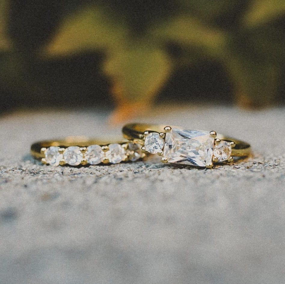 A gold engagement ring with a rectangular stone and side diamonds, set next to a matching diamond-studded wedding band.