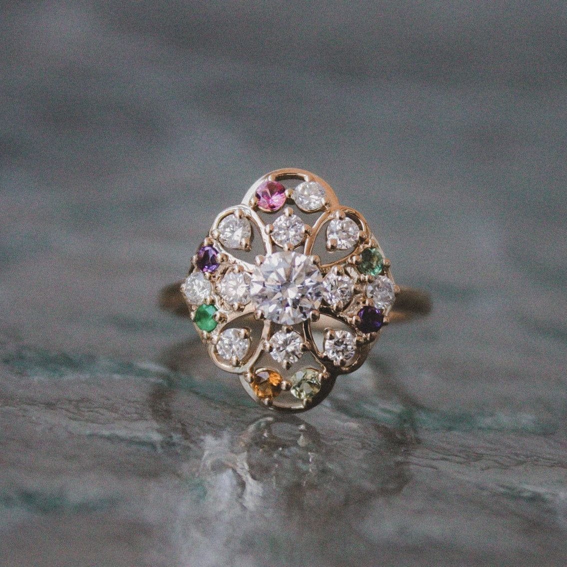 A gold ring with a central diamond surrounded by a floral arrangement of smaller diamonds and colored gemstones.