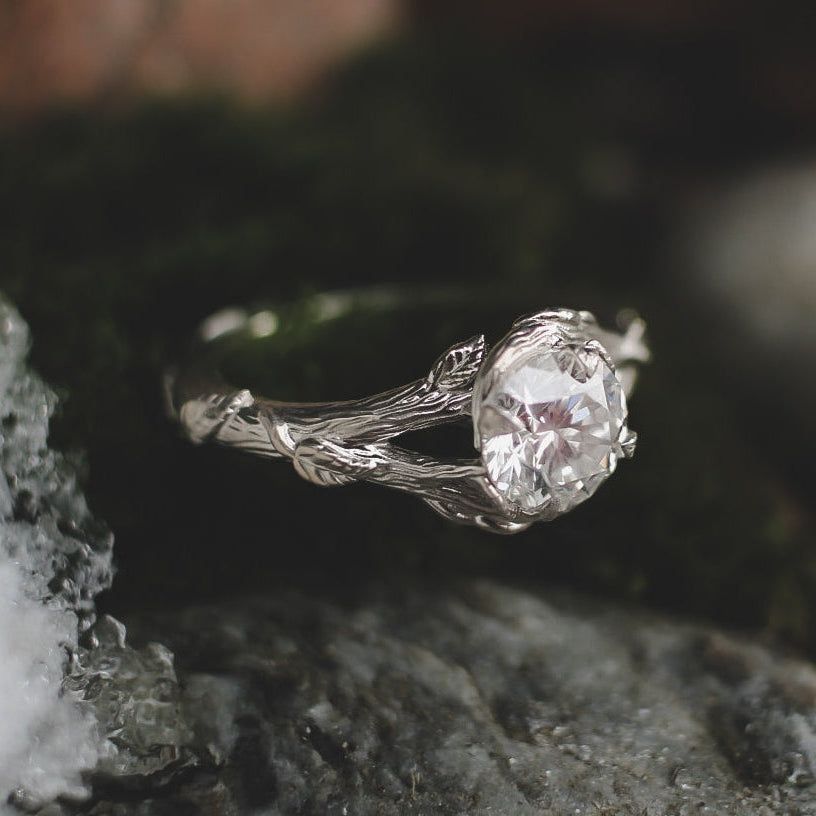 A silver engagement ring with a branch-like, textured band and a round center stone, resting on mossy, dark stone.