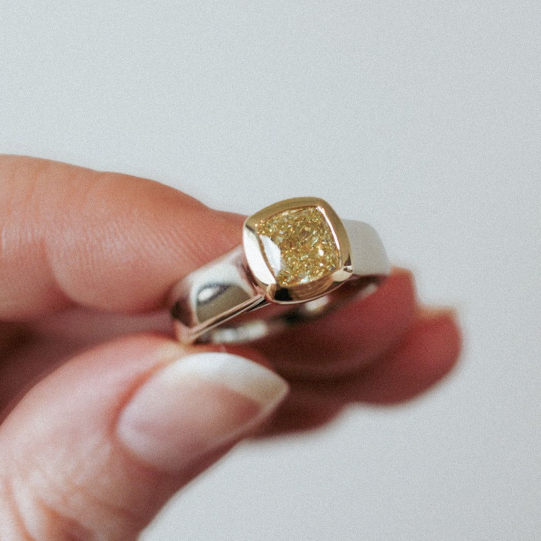 A hand holds up a silver ring with a square, textured gold centerpiece.