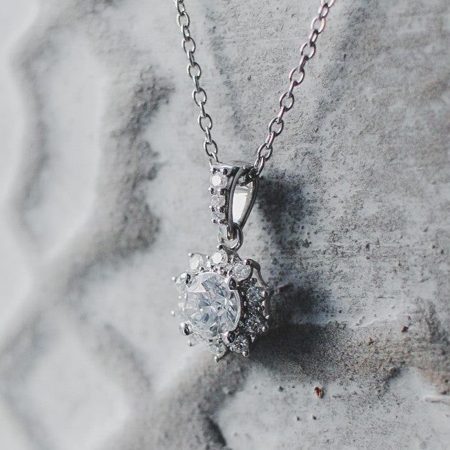 A silver pendant necklace featuring a round central stone surrounded by smaller crystals against a textured gray background.