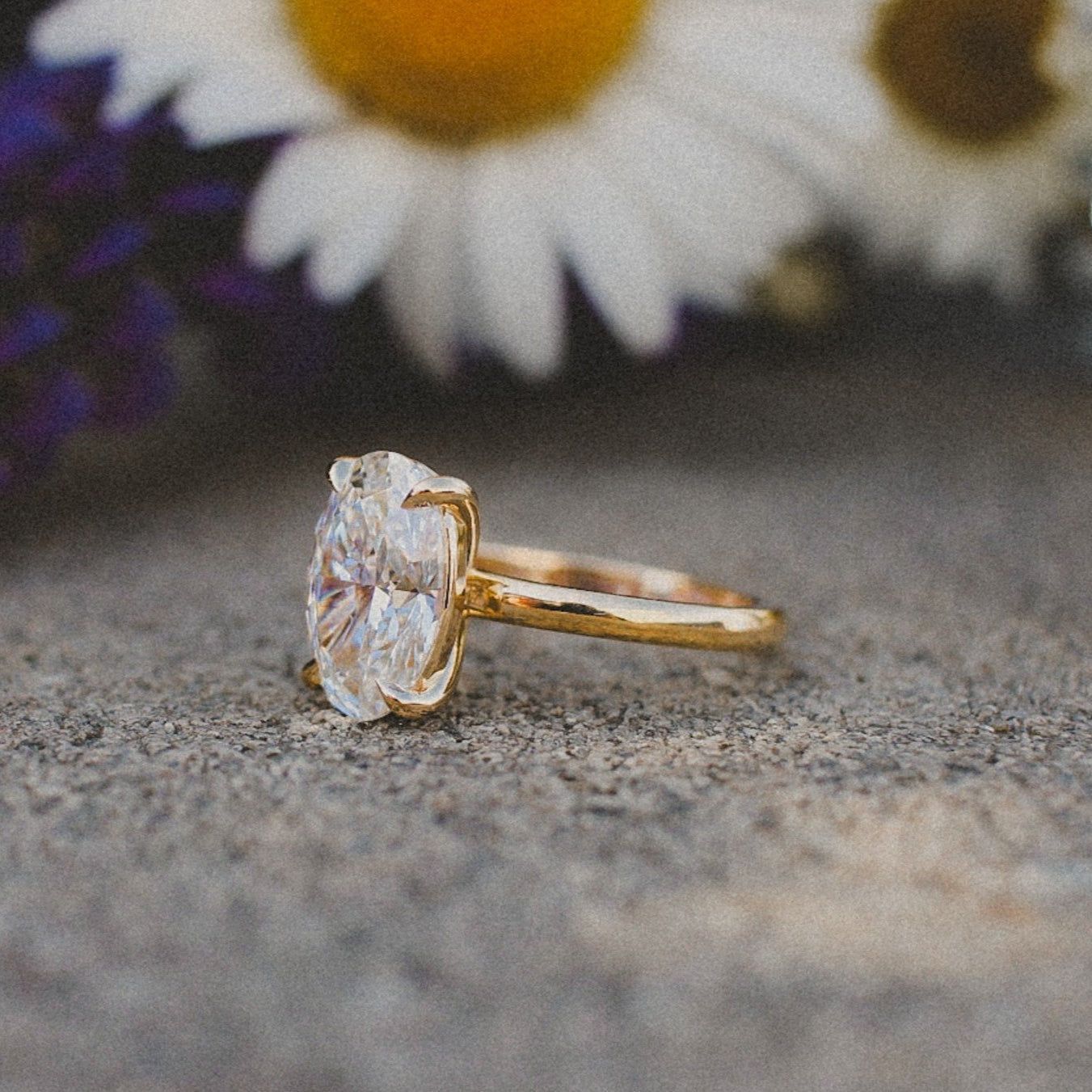 An oval-cut diamond engagement ring with a gold band rests on a stone surface in front of white daisy flowers.