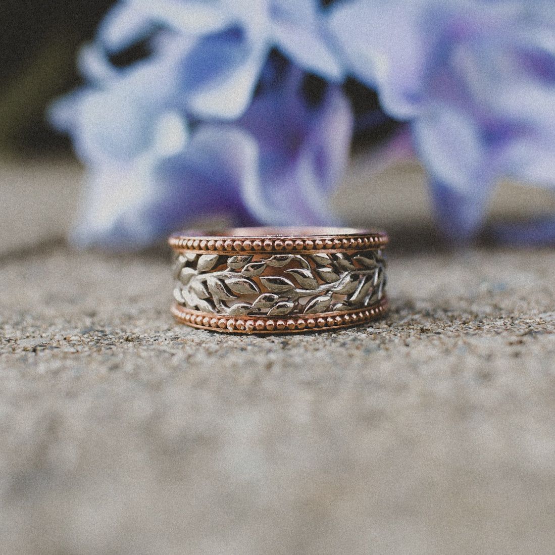 A two-toned ring with a silver leaf-patterned band bordered by beaded rose gold, resting on a stone surface.