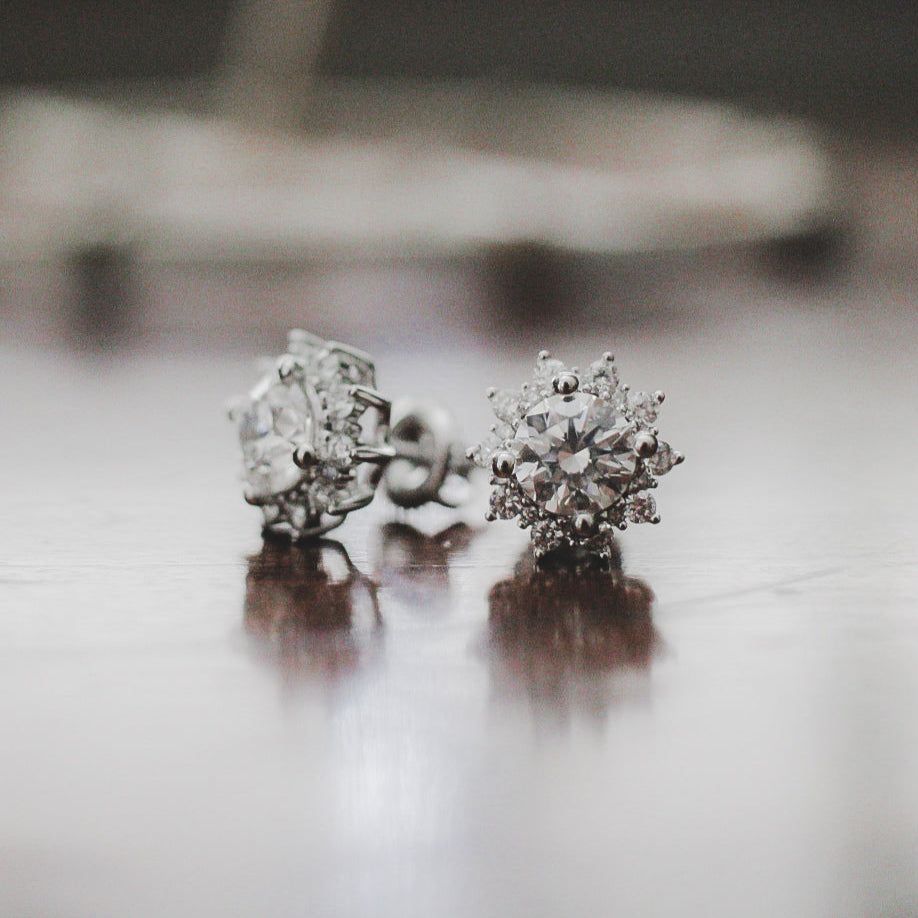 Two silver stud earrings with large center stones surrounded by a halo of smaller, sparkling stones on a dark surface.