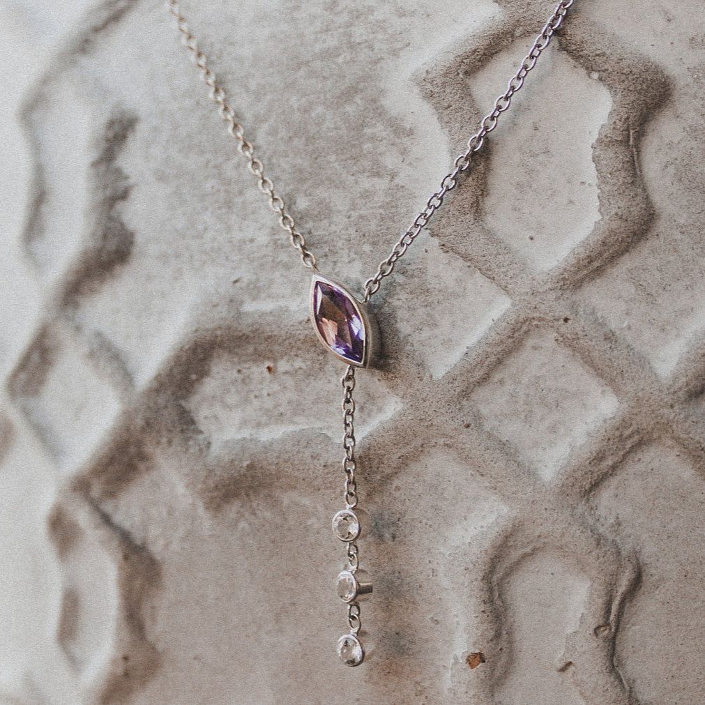 A delicate silver lariat necklace with a central purple marquise-cut gem and three small dangling clear stones.