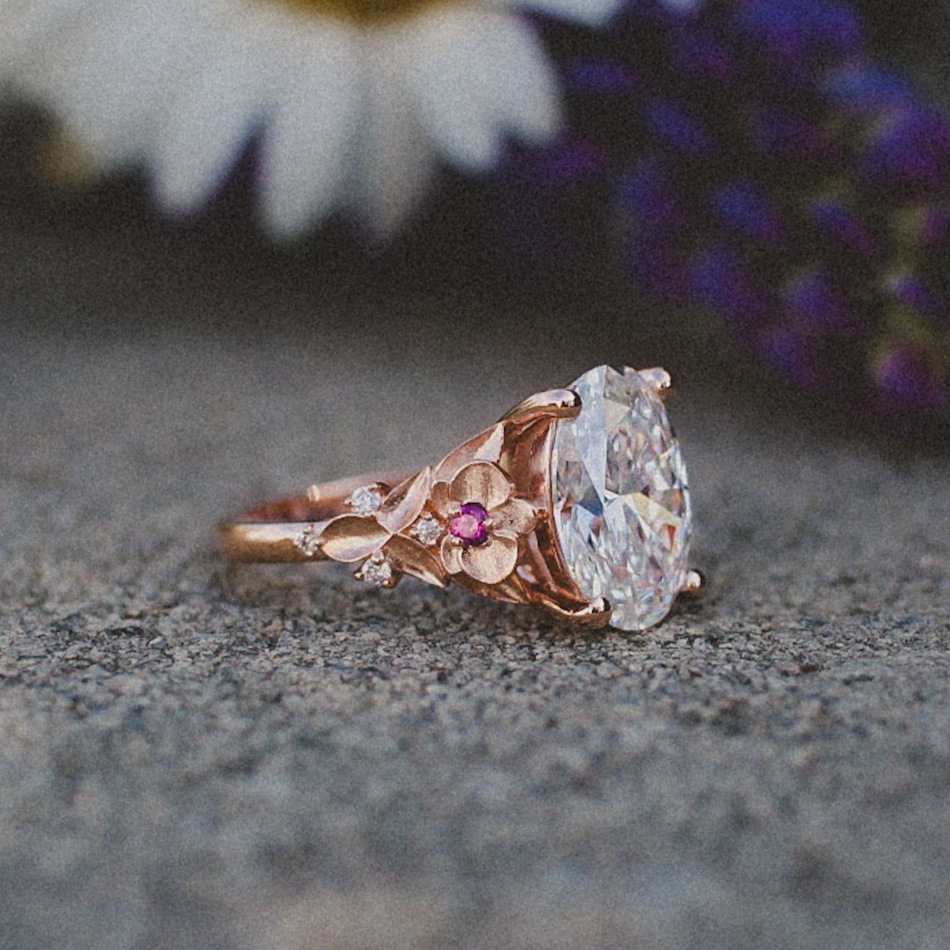 An oval-cut diamond ring in rose gold with floral detailing and a small pink accent stone, set on a textured surface.