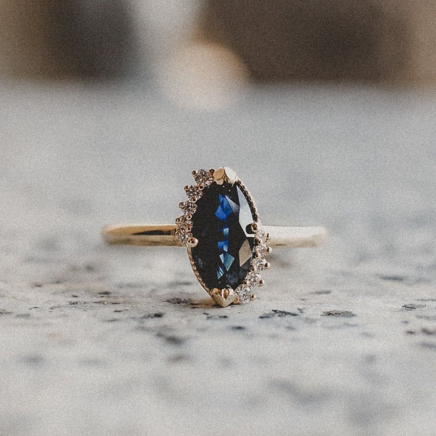 A gold ring with a marquise-cut dark blue sapphire, accented by a row of small diamonds on one side, on a stone surface.