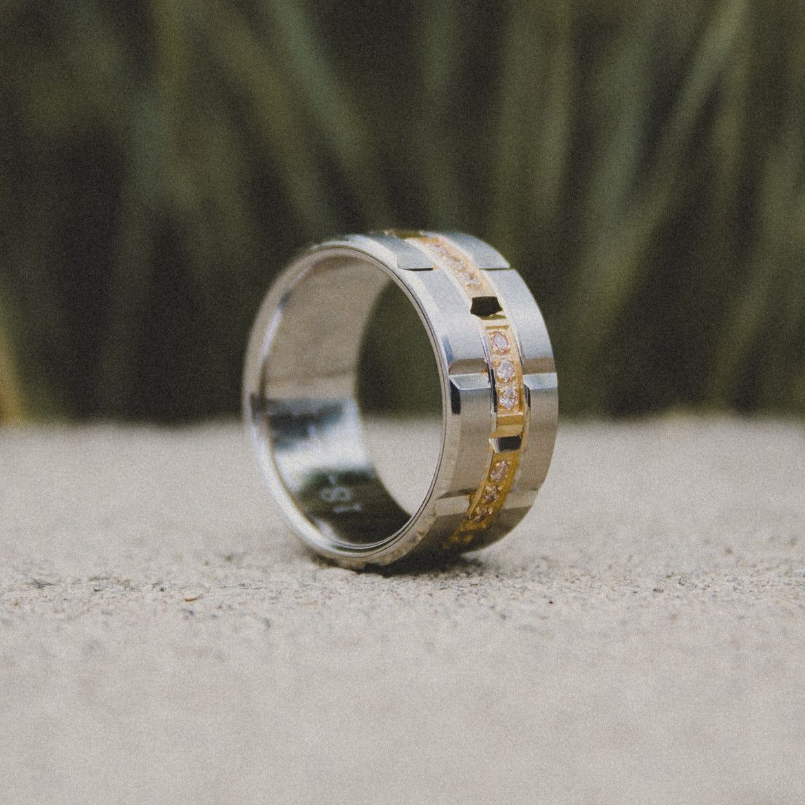 A two-tone metal wedding band with a central inlaid gold channel set with small diamonds, resting on a textured surface.