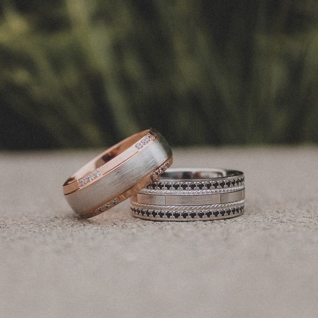 Two wedding bands rest on a textured surface, one featuring rose gold and brushed silver, the other silver with black detail.