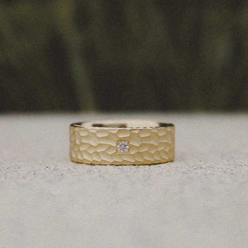 A hammered gold ring with a single small, round, inset diamond centered on a textured, matte surface.