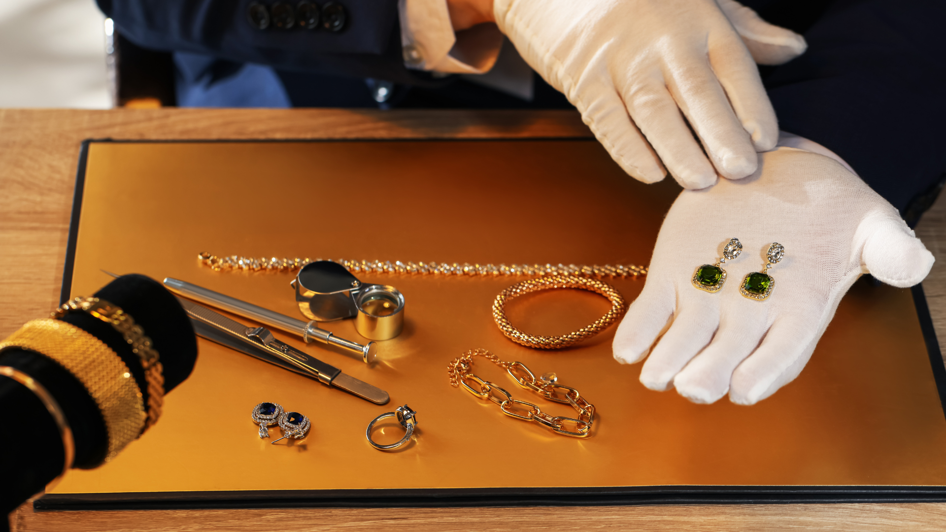 A person wearing white gloves displays a pair of green gemstone earrings on a golden tray filled with assorted jewelry.