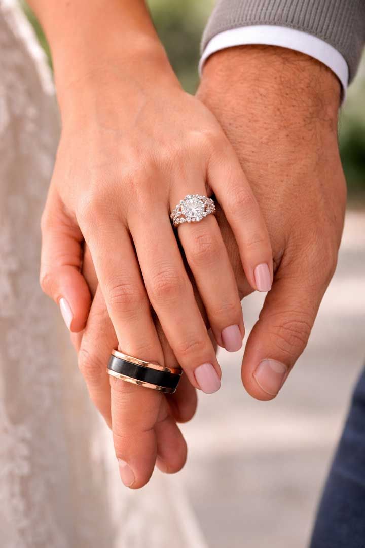 Two hands held together, showcasing a diamond engagement ring on one hand and a black and gold band on the other.