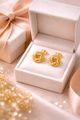 A pair of gold knot-style stud earrings rests inside an open, cream-colored jewelry box next to a gift box with a ribbon.