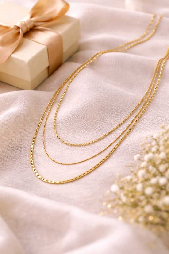 A multi-layered gold chain necklace rests on soft, light pink fabric next to a gift box with a ribbon and flowers.